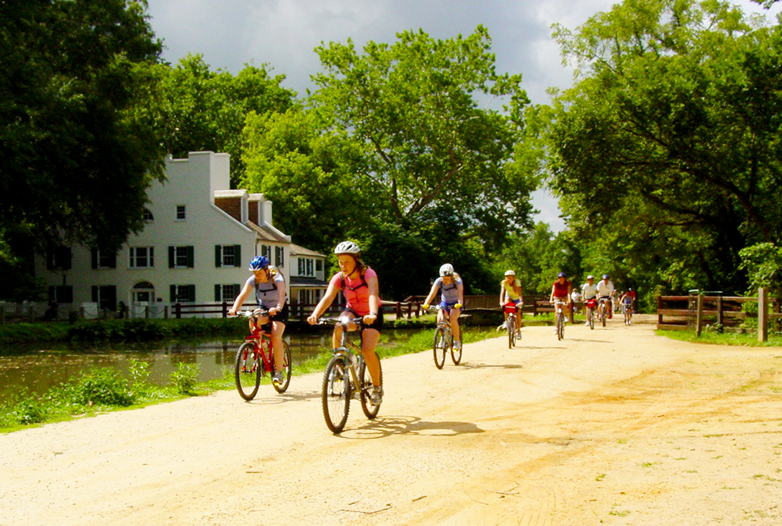 A group of young people ride their bikes along a wide dirt path past an old white building and a waterway bordered by trees.