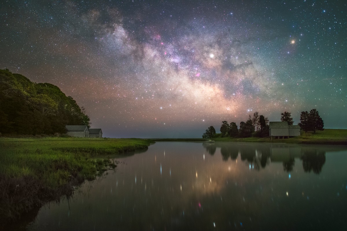 The Milky Way Galaxy shines brightly with thousands of stars above a reflective pond with grassy green banks and small stone buildings rising in the background with dark leafy trees.