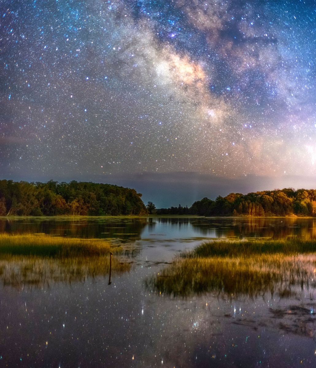 Green and orange trees stand along the water, reflecting the bright and starry light blue and purple sky above.