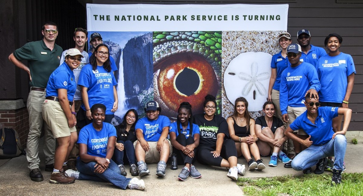 A group of young volunteers in matching NPS t-shirts pose around a large, creative Centennial sign.