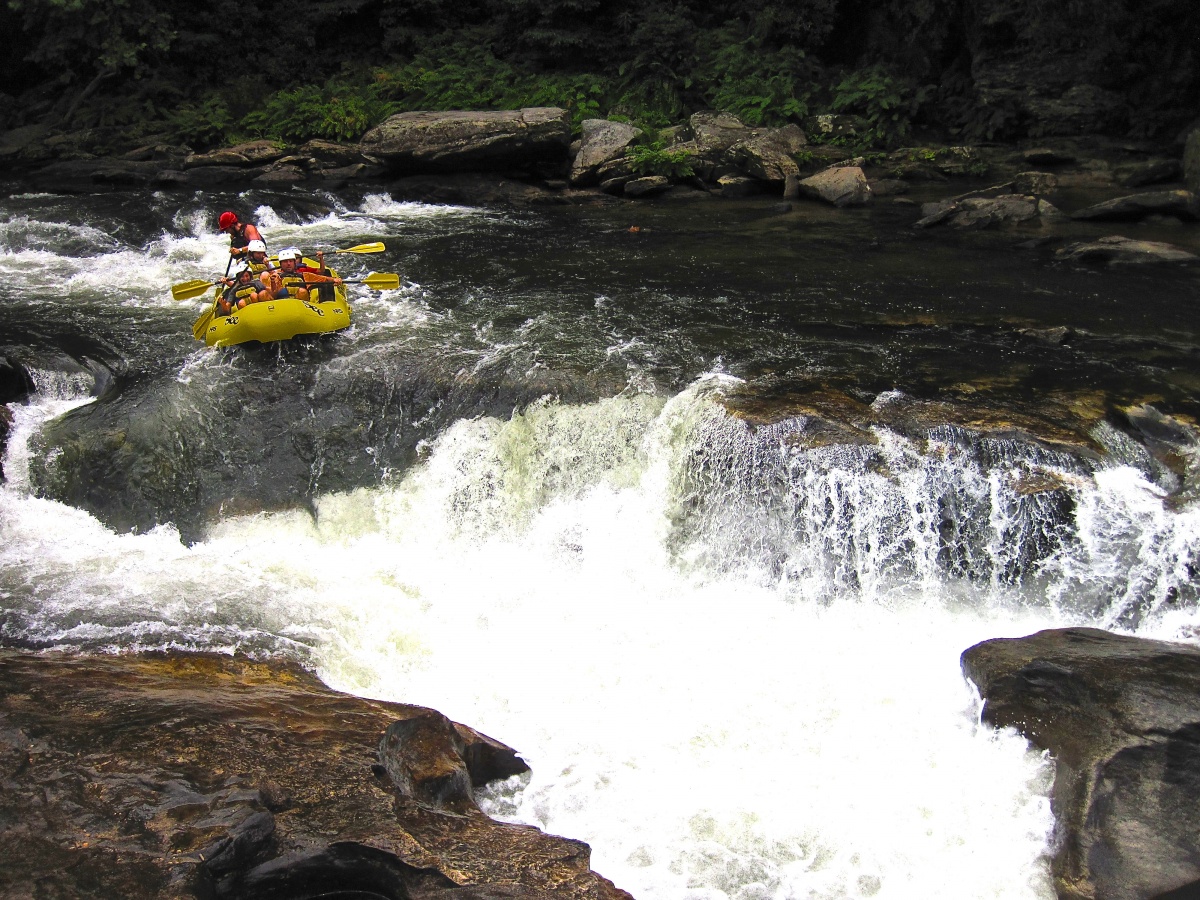 Five people with helmets sit in a yellow rubber boat and paddle down a river towards a large whitewater rapid and rocks.