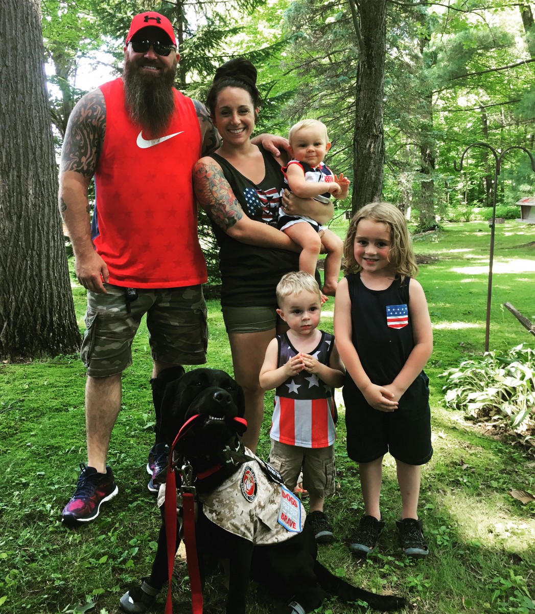 A beaming husband and wife with their three children and family dog pose in a sunny, green park.