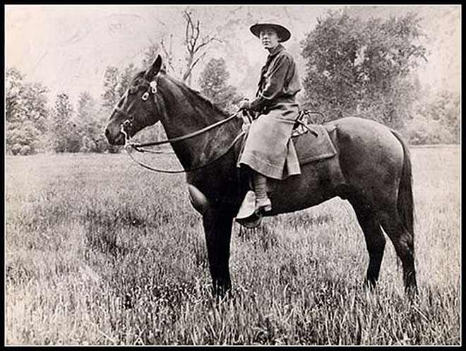 A woman in a riding skirt and ranger's hat sits on horseback among brush and in front of blurred trees and mountains.