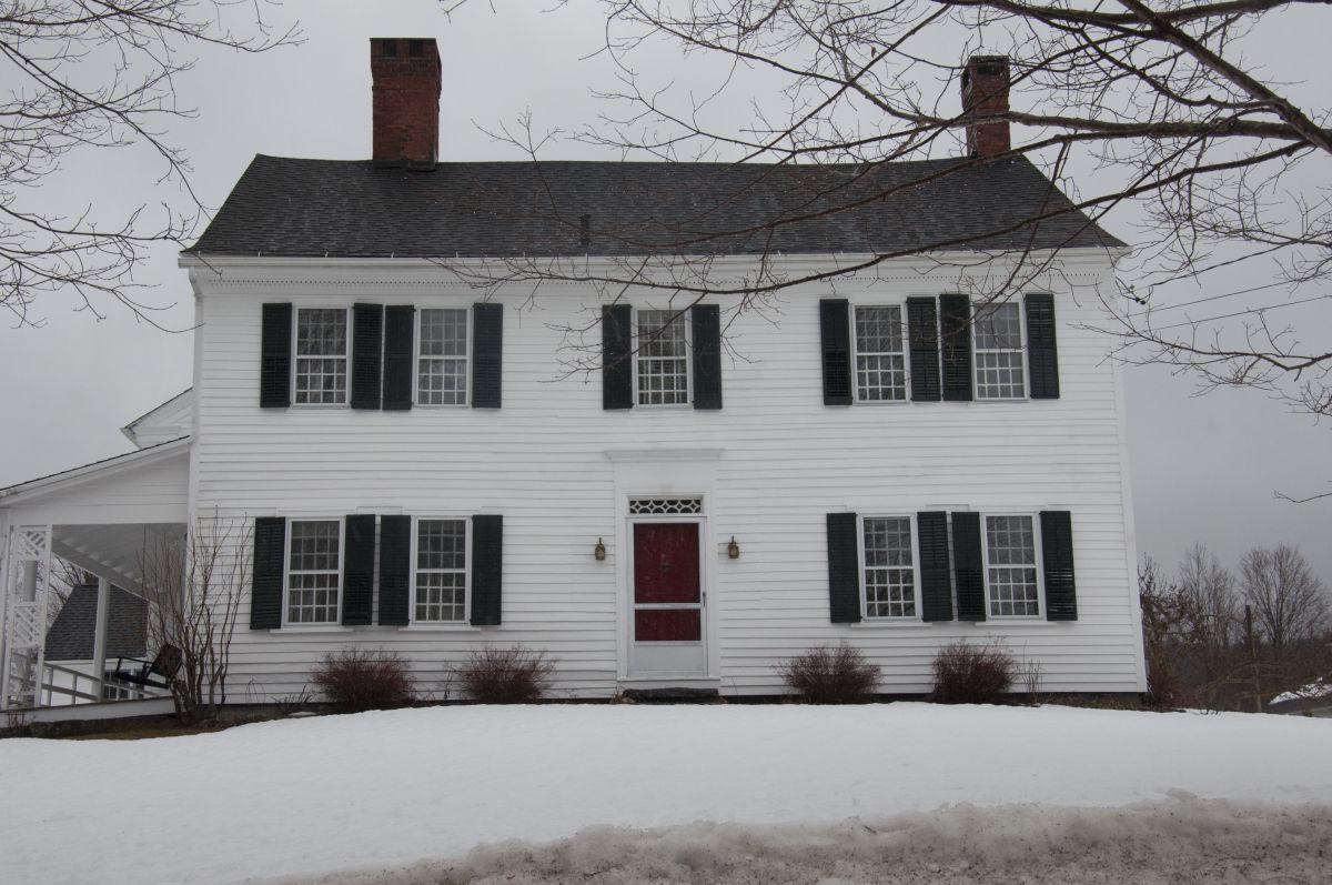 A white building with black shutters and a red door stands alone in the snow.