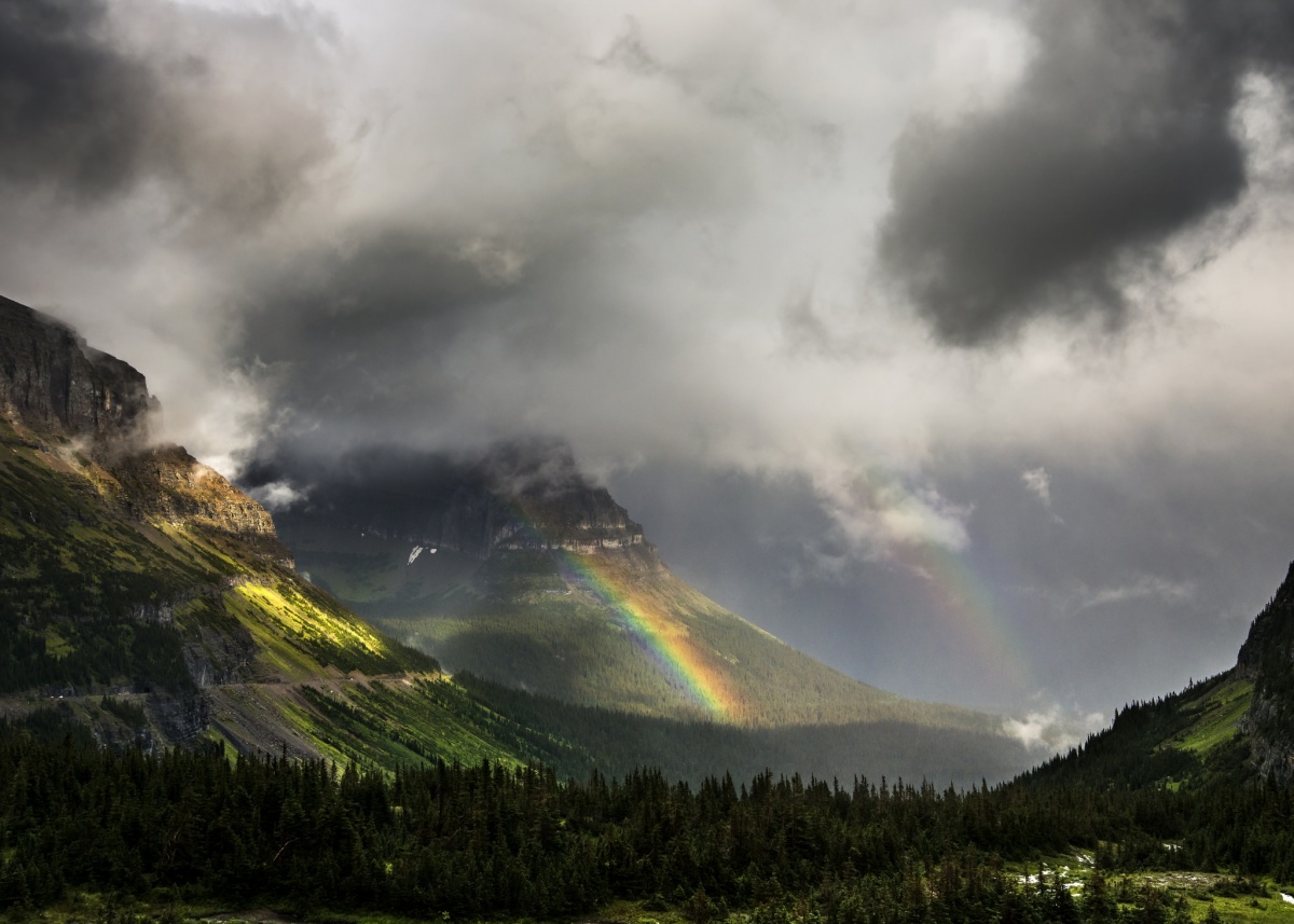 With cloudy skies overhead, a rainbow emerges over green covered mountains and forests.
