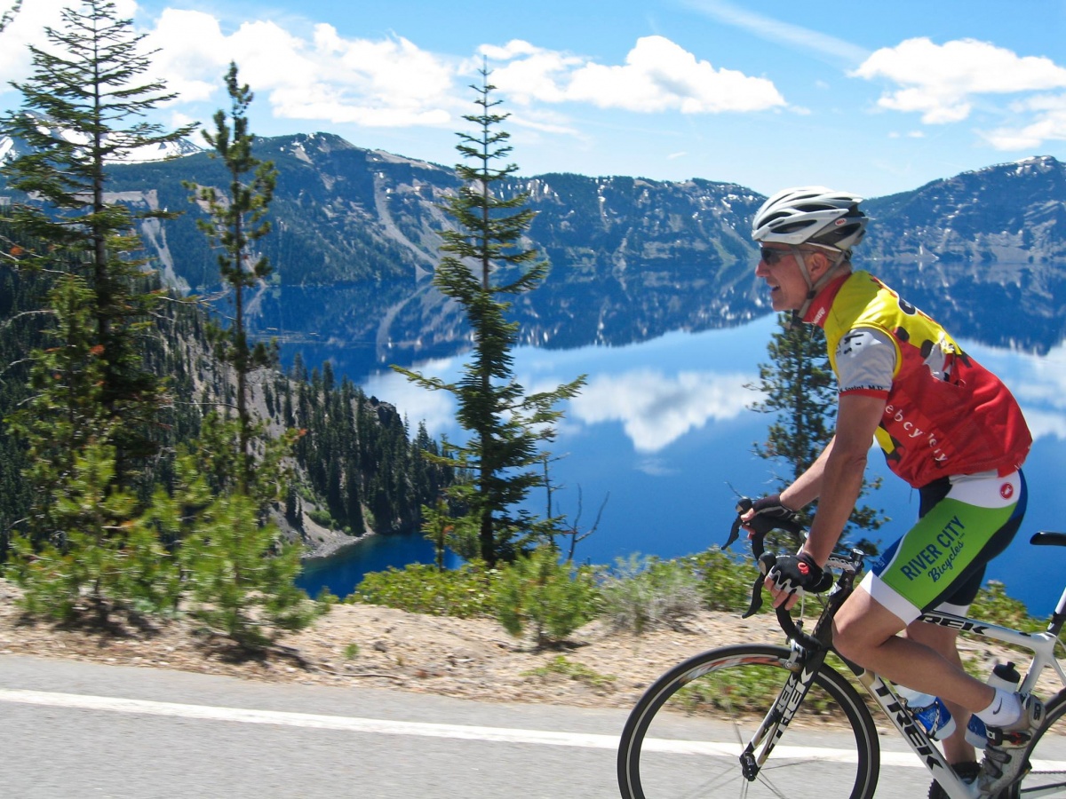 um homem branco magro e mais velho usando um capacete e roupas de ciclismo brilhantes monta uma bicicleta em uma estrada que passa por um lago blue mountain.ciclismo em torno de Crater Lake é divertido! Foto do National Park Service.