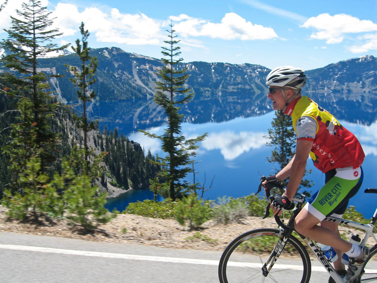 A white man in tight biking clothes and a helmet rides a bike past a blue mountain lake.