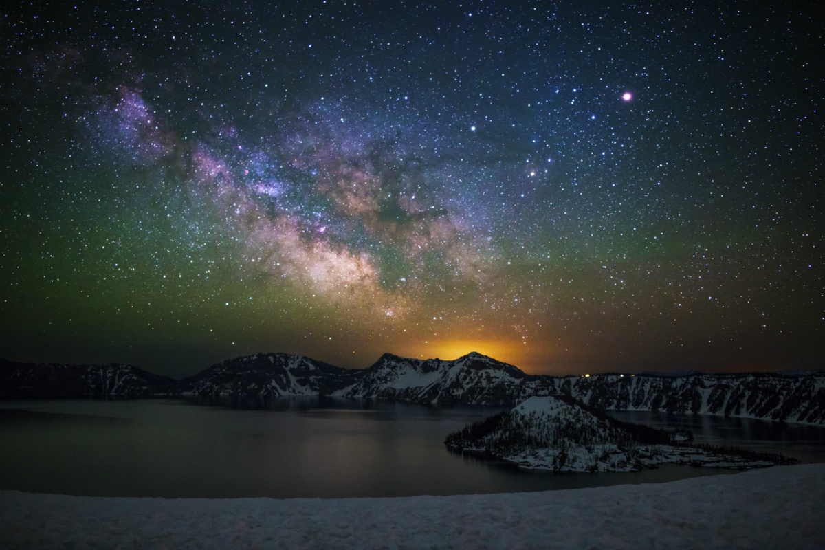 Un deslumbrante cielo nocturno de estrellas y la vía láctea brilla sobre acantilados cubiertos de nieve que descienden hasta un amplio lago circular.