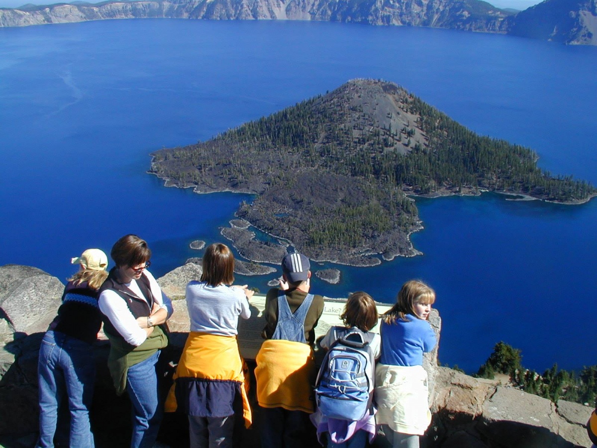 En hvit kvinne og fem hvite barn står ved en fjellvegg med utsikt over en øy i en bred blå innsjø.En naturskjønn utsikt tilbyr en fantastisk utsikt Over Wizard Island. Foto Av National Park Service.