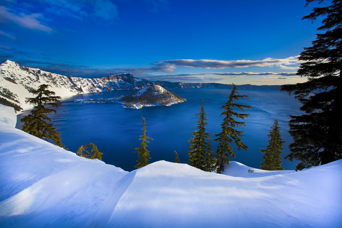High snow covered cliffs surround a large, round blue lake with a small cone shaped island in the middle of it.