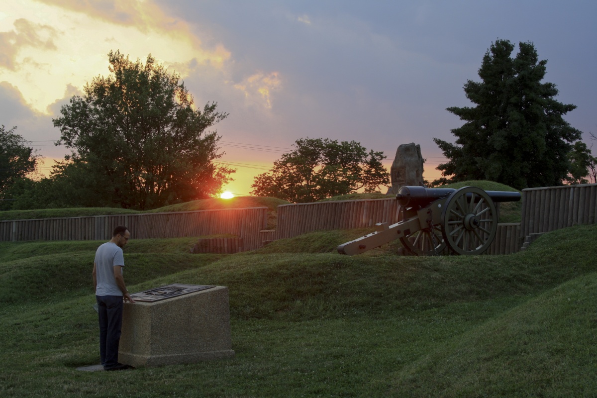 An orange sunrise peeks over green hills. A man reads a plaque in front of a cannon.