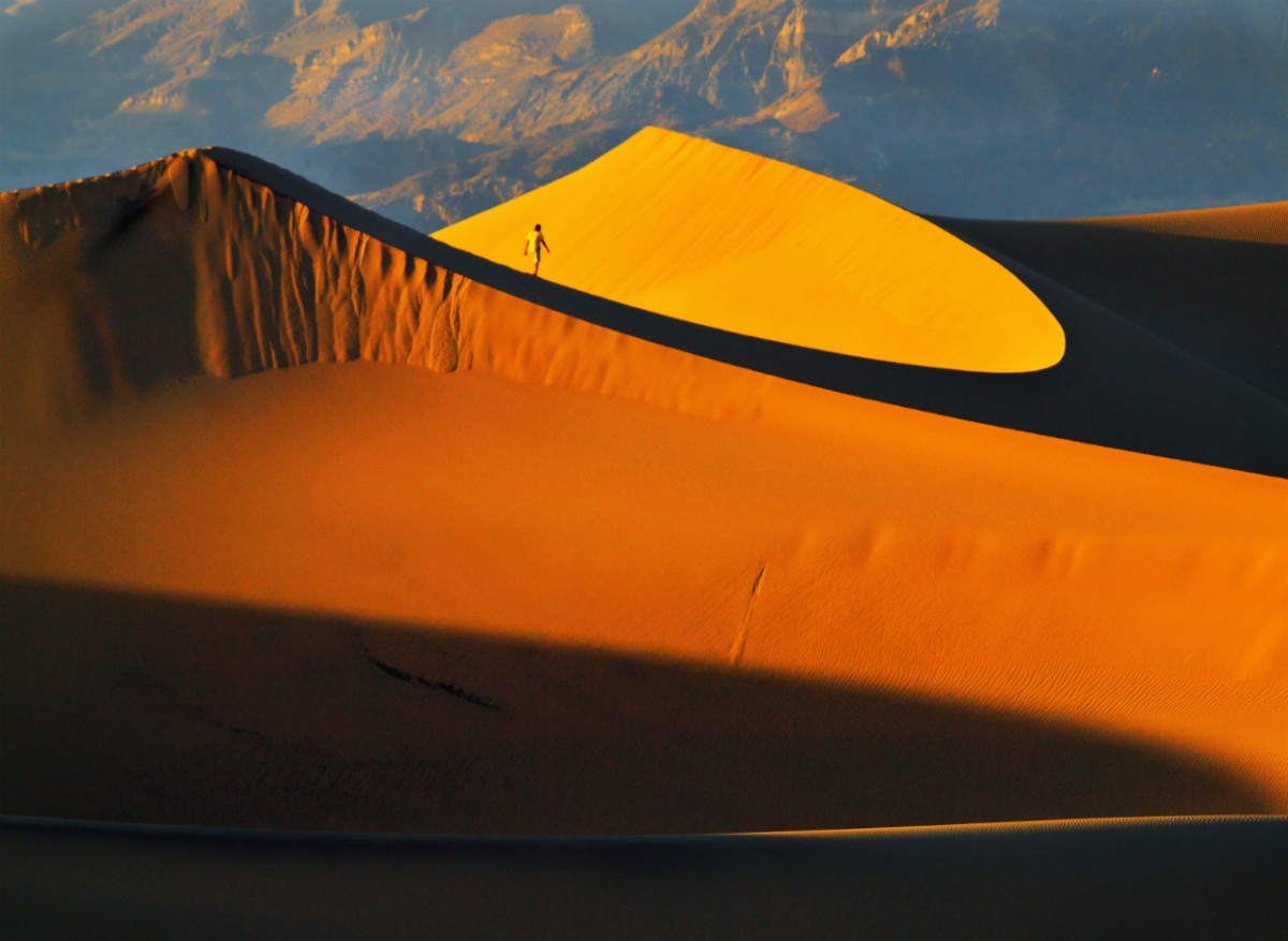 A lone man walks on the ridge of a high sand dune in the golden light of sunset.