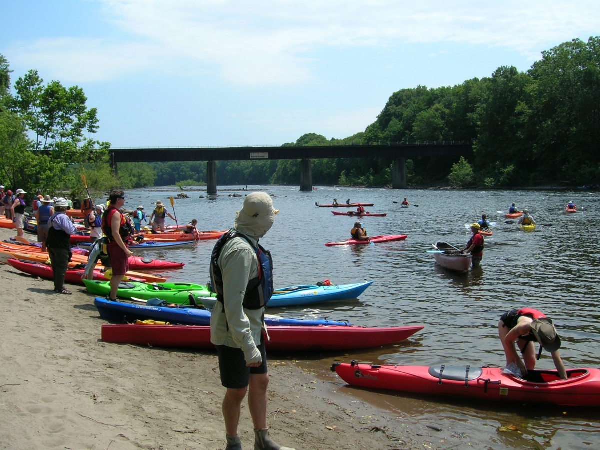 A large crowd of people with dozens of colorful kayaks stand on the bank of a wide, calm river with some kayaks in the water paddling downstream past a highway bridge.