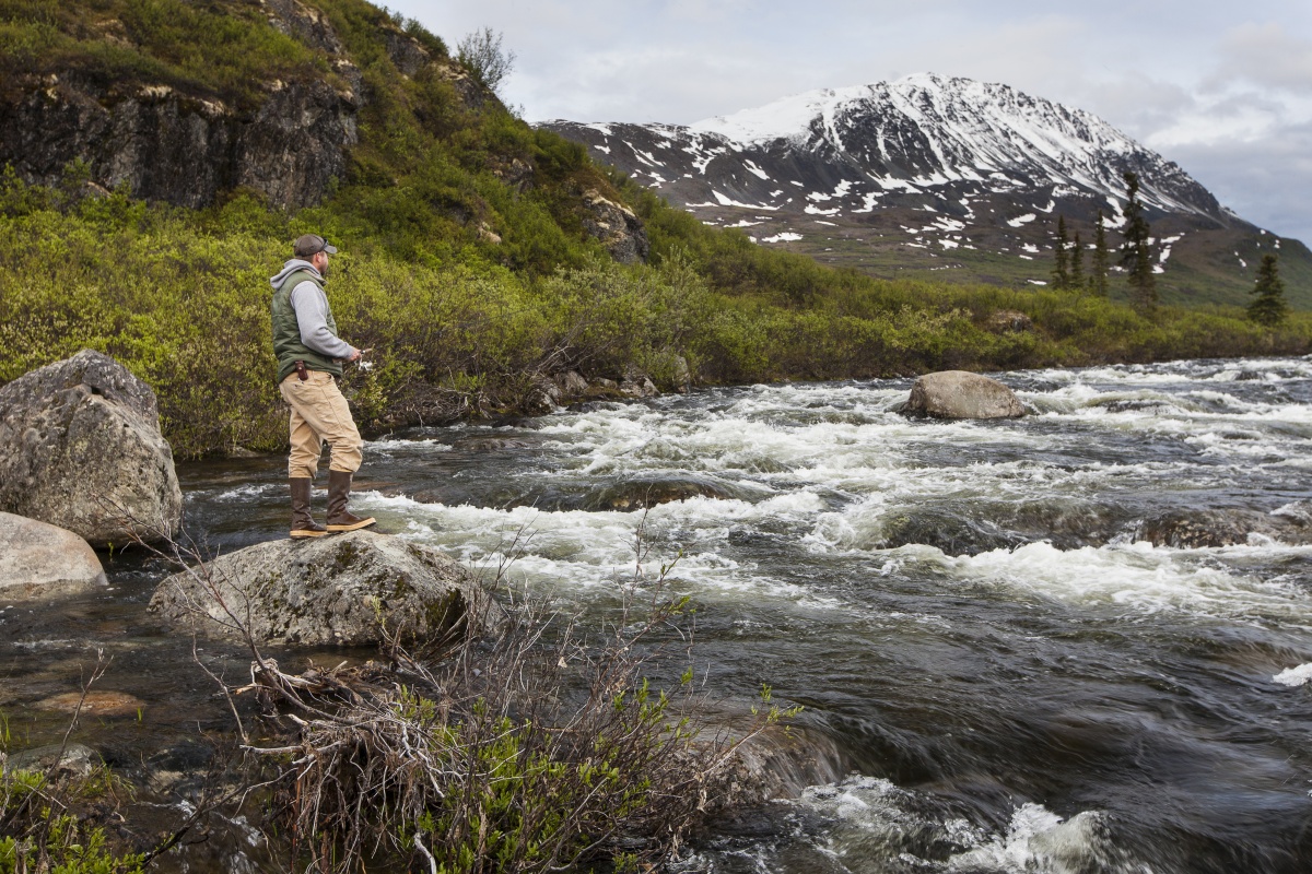 A man wearing outdoor gear and fishing with a rod and reel stands on a rock on the bank of a rushing river.