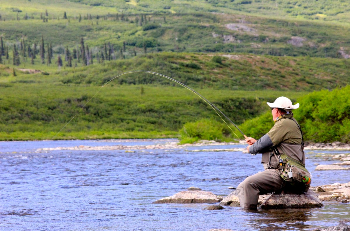 A man sits on a rock along a scenic flowing river as rolling hills carpeted in green bushes and trees rise in the background.