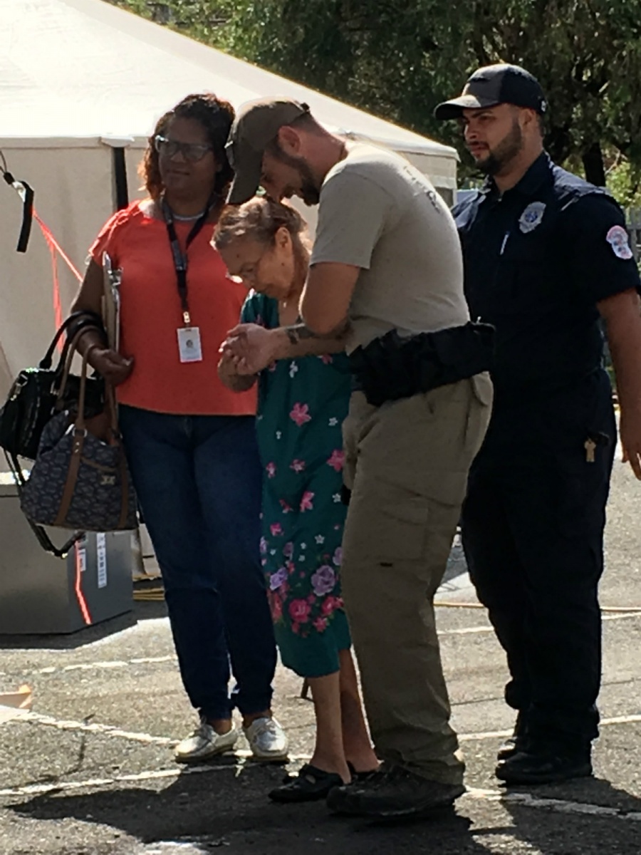 Two policemen and a woman help an elderly woman walk across a parking lot to a tent.