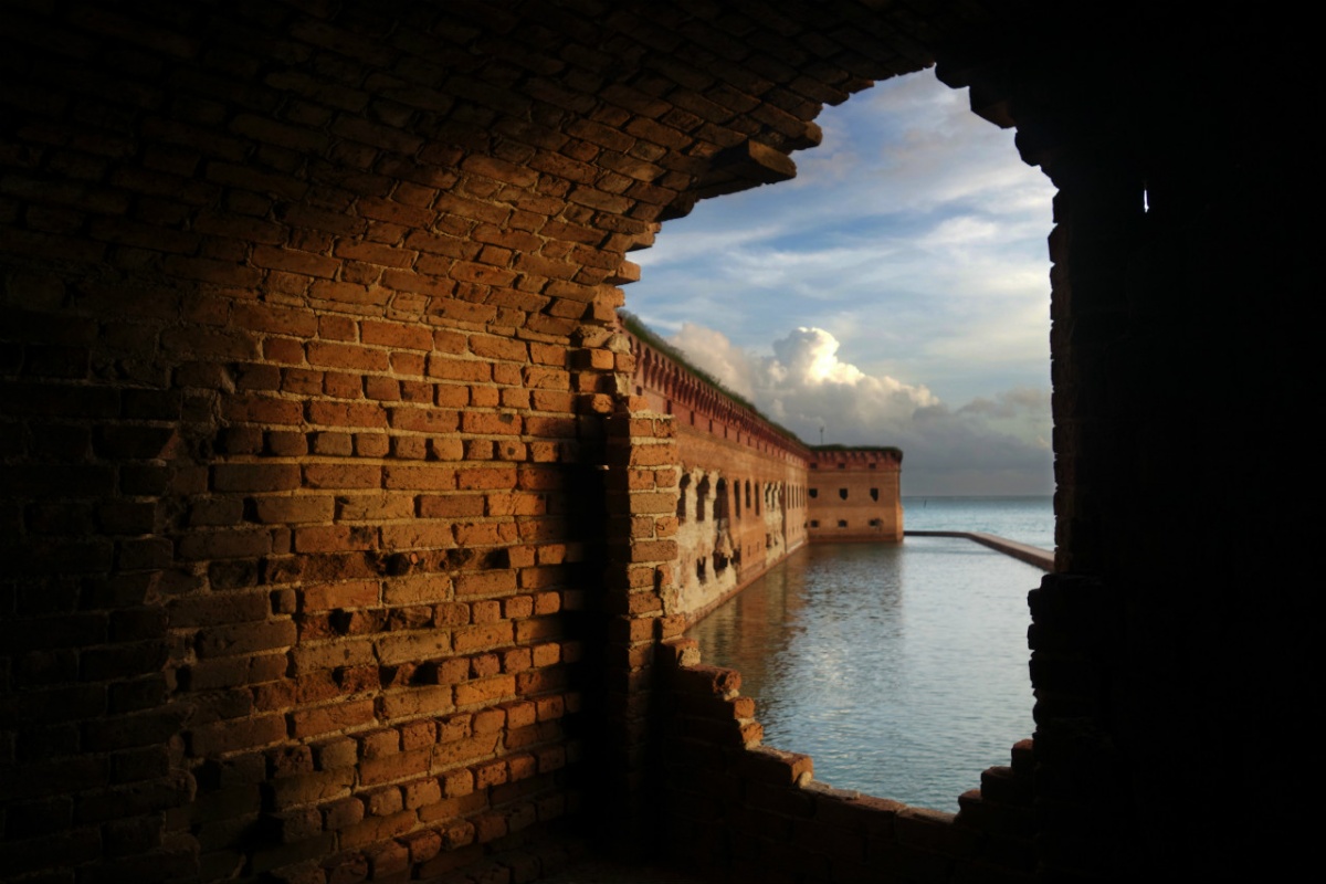 A hole in a red brick wall allows for the dark blue water and cloudy blue sky outside to be seen. 