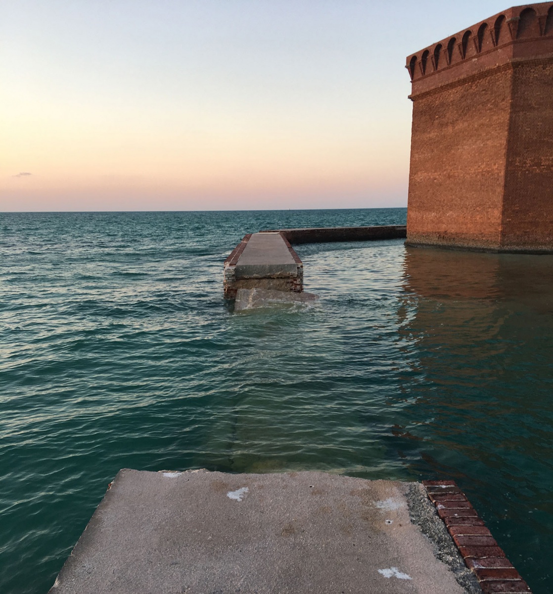 A concrete and brick walkway shows a large gap where a part of it collapsed into shallow blue water.