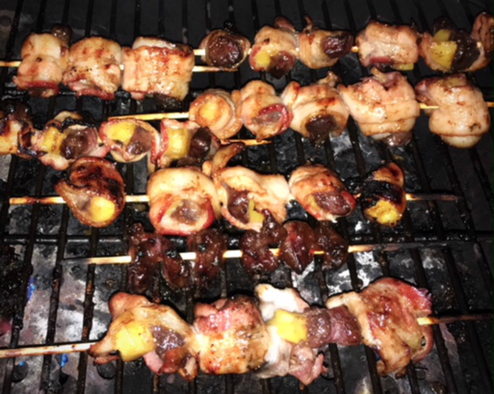 Seven skewers filled with cubed chicken, pineapple chunks, and bacon are slightly charred and sitting on a grill.