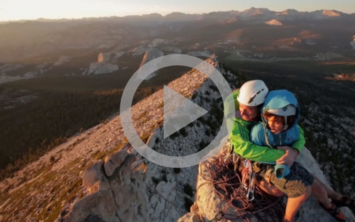A parent and child wearing mountaineering gear hug on top of a mountain with a pretty landscape in the background.