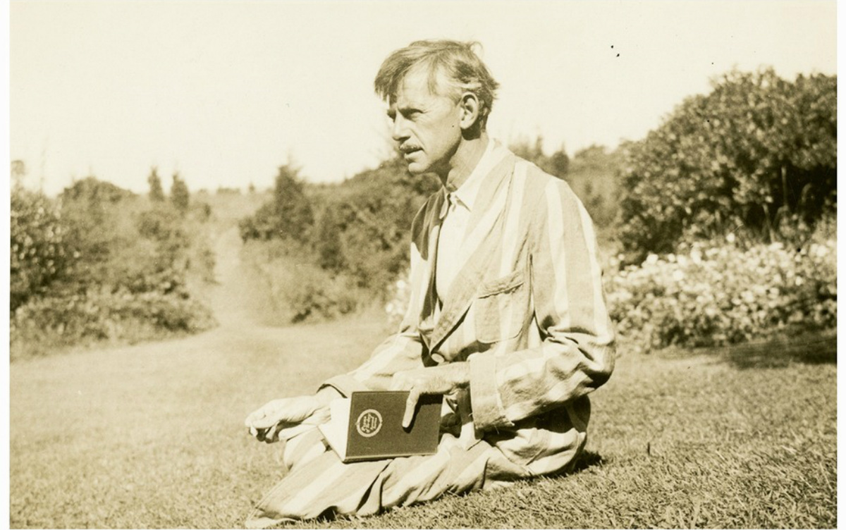 Eugene O’Neill, aging with squinted eyes and a graying mustache, sits in a striped bathrobe with a book on the grass in a vineyard in a yellowed historical photograph.