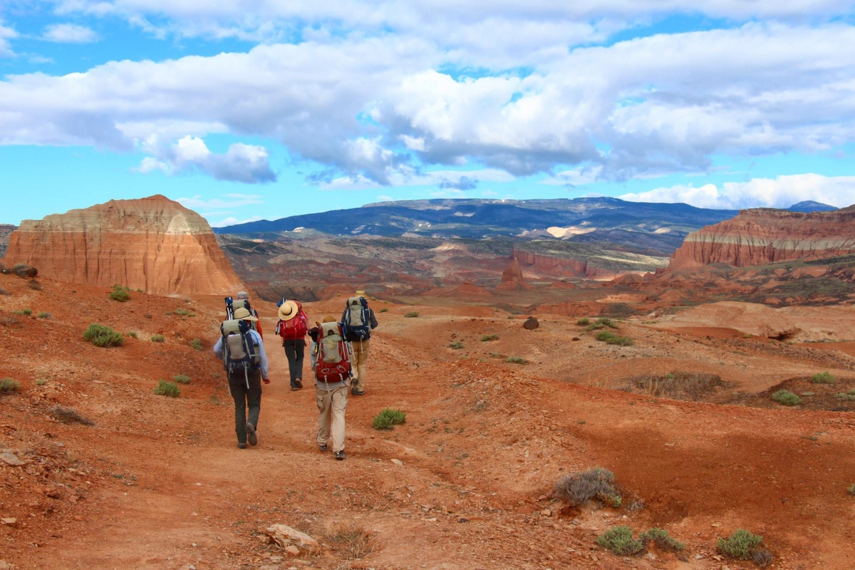 Five people with backpacks hike down a dirt path on a rocky desert plain.