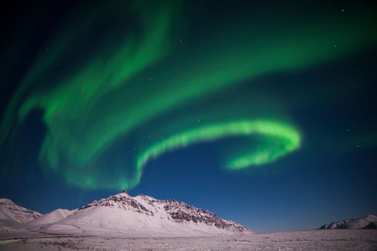 Three long lines of bright green light curve across the night sky above a snow covered landscape studded with small mountains.