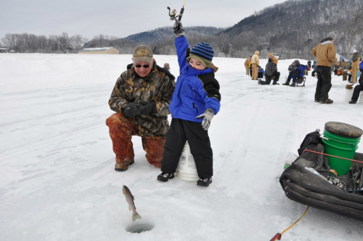 Standing on a frozen lake, a boy uses a fishing rod to pull a fish out of a hole in the ice with an older man helping him.