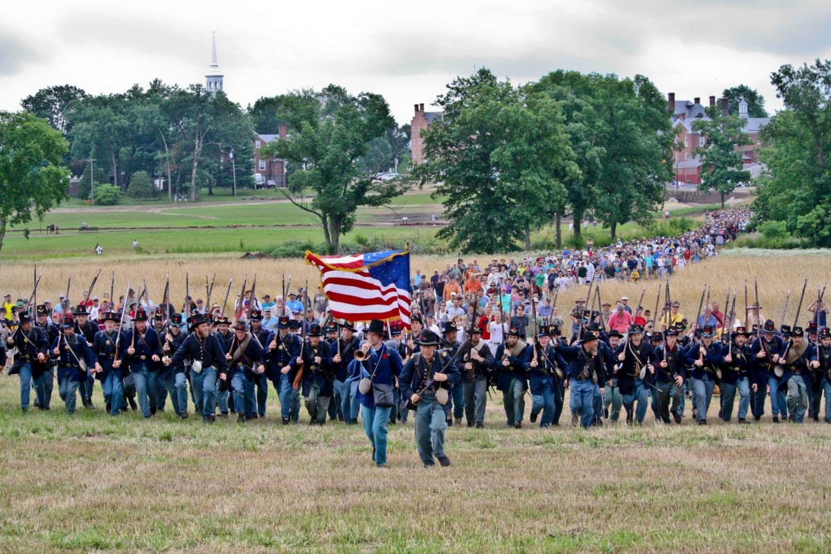 A group of men dressed in soldier's uniforms lead a large group of people to a field. The men carry a large red, white and blue flag.