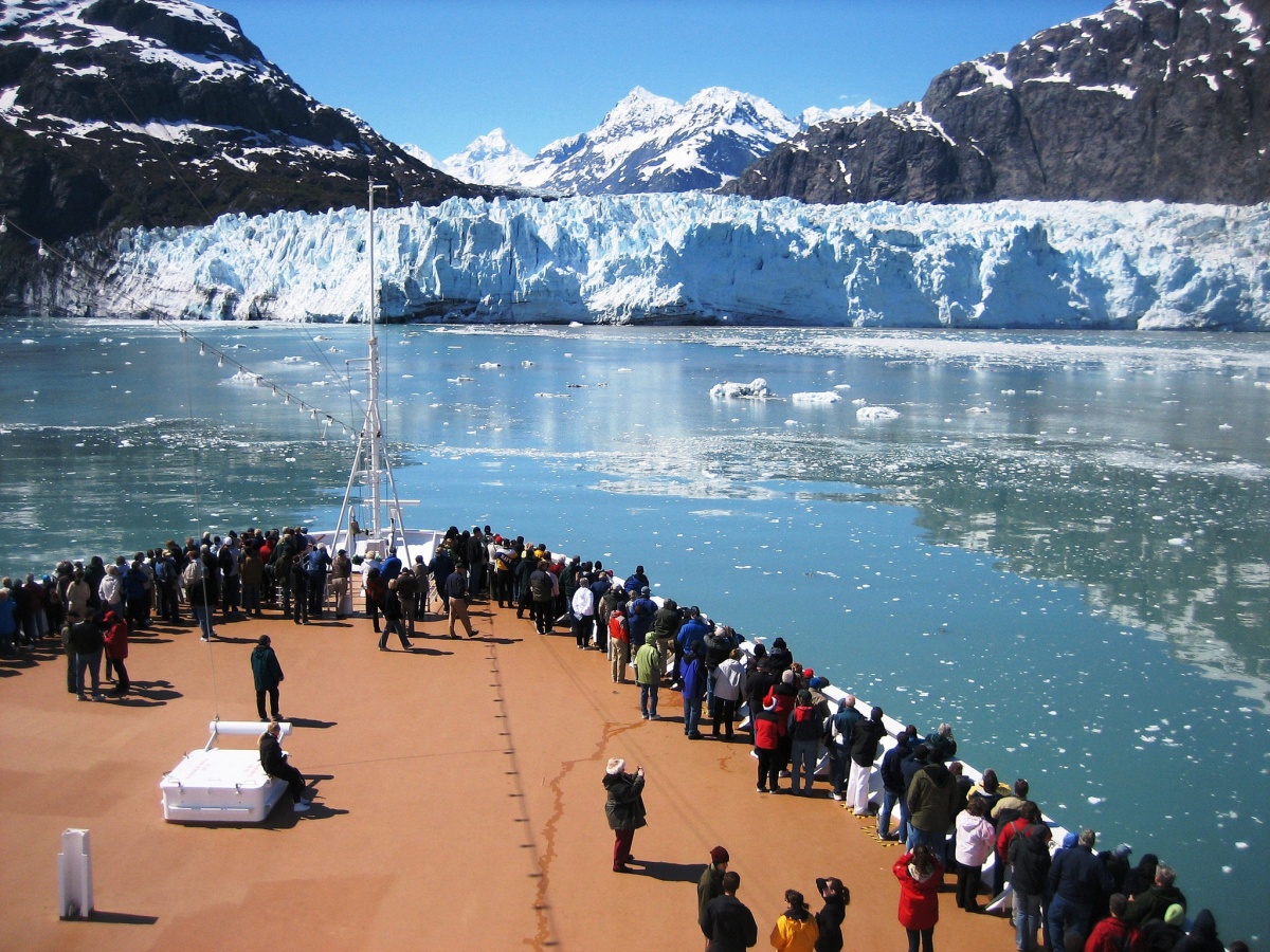 A large group of people stand on the deck of a ship looking across a bay where a glacier runs down from a mountain into the water.