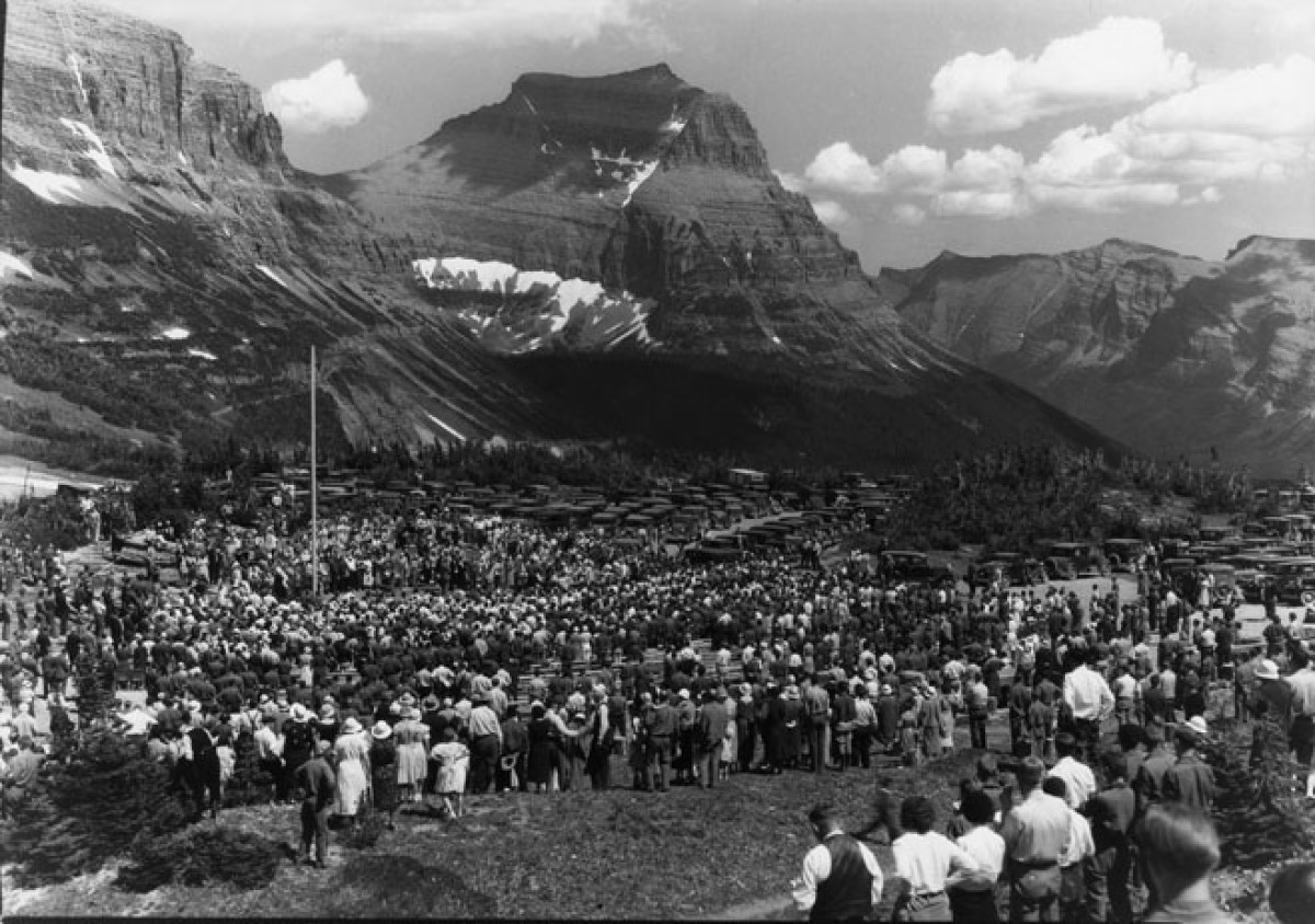 Massive crowds line the grassy hills and face inward toward a lone flagpole with a range of tall mountains and puffy clouds rising in the background.