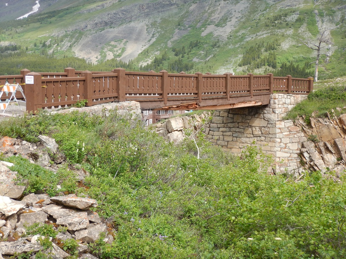 A wooden pedestrian bridge shows major signs of damage to its middle span as it crosses a wide ditch in a mountain valley.