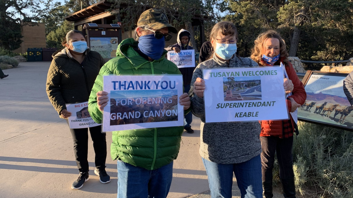 A small group of people wearing masks stand in a courtyard holding signs that say "Thank You for Re-opening the Grand Canyon."