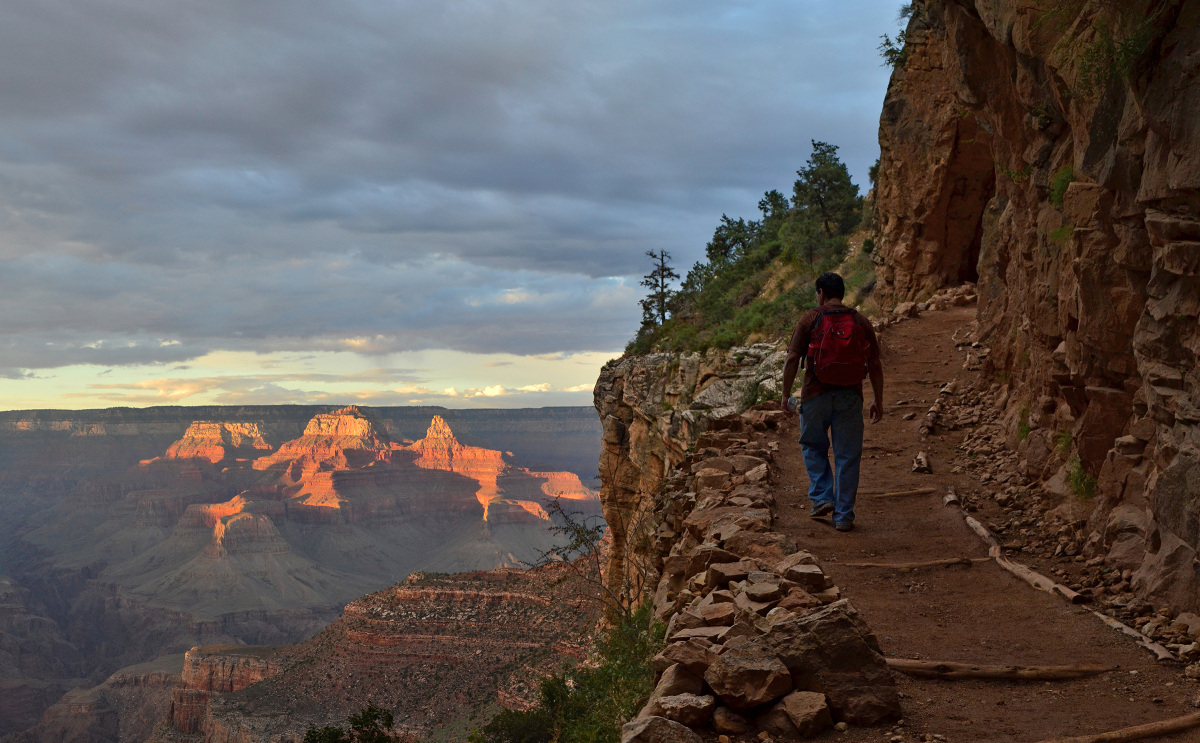A man wearing a backpack walks along a dirt trail overlooking a massive canyon.