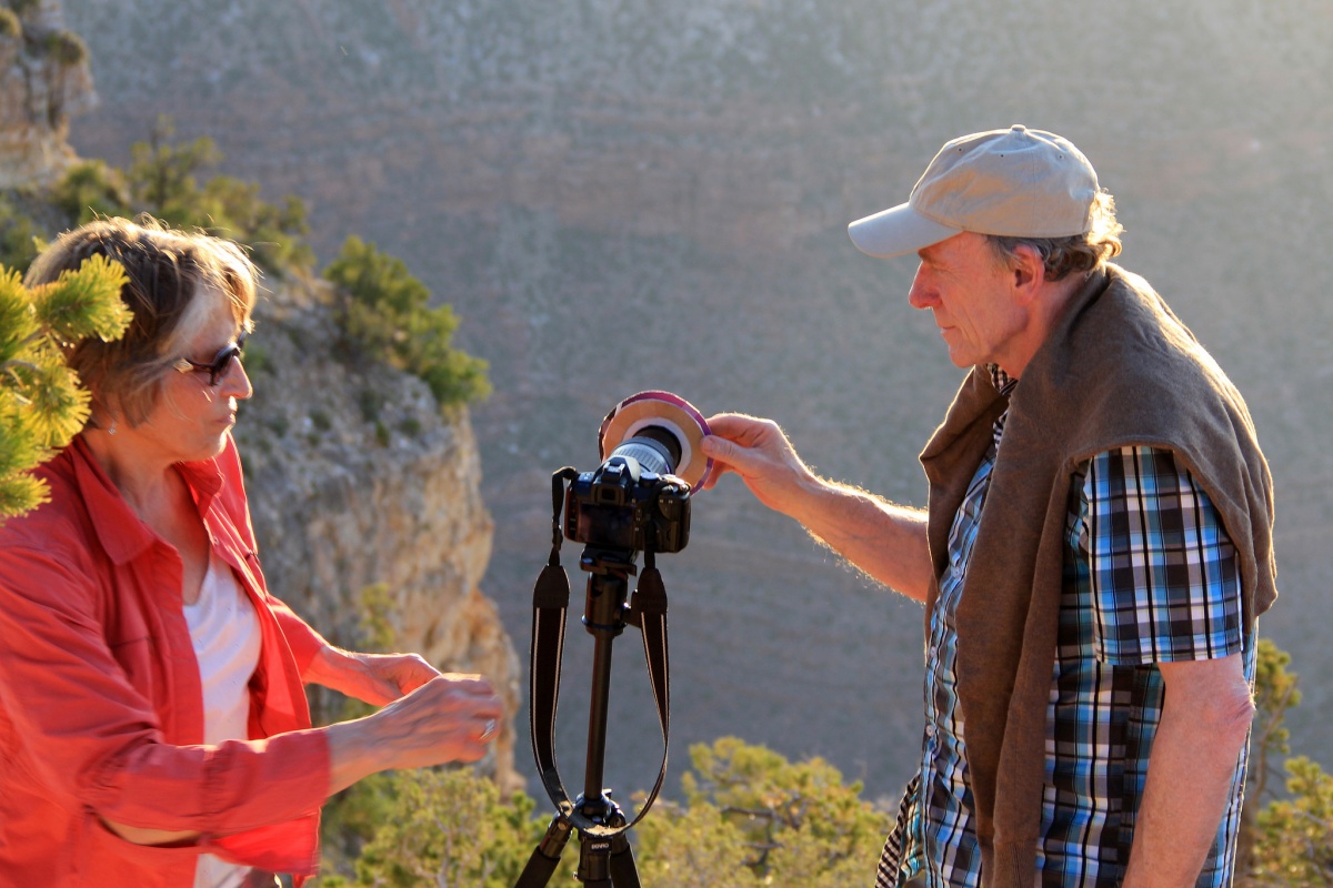 A man and a woman make adjustments to a camera on a tripod while looking out over the cliff wall of a massive canyon.