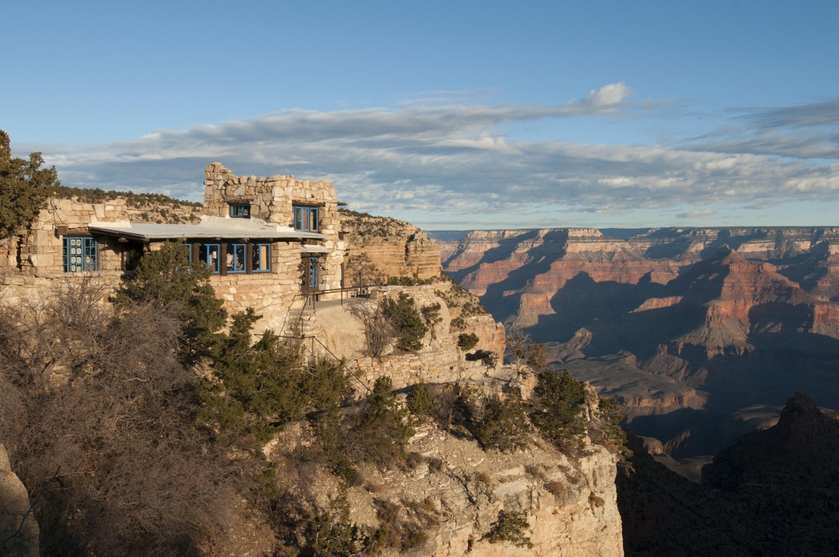 A stone building with blue window frames stands on the edge of a cliff overlooking a massive canyon.