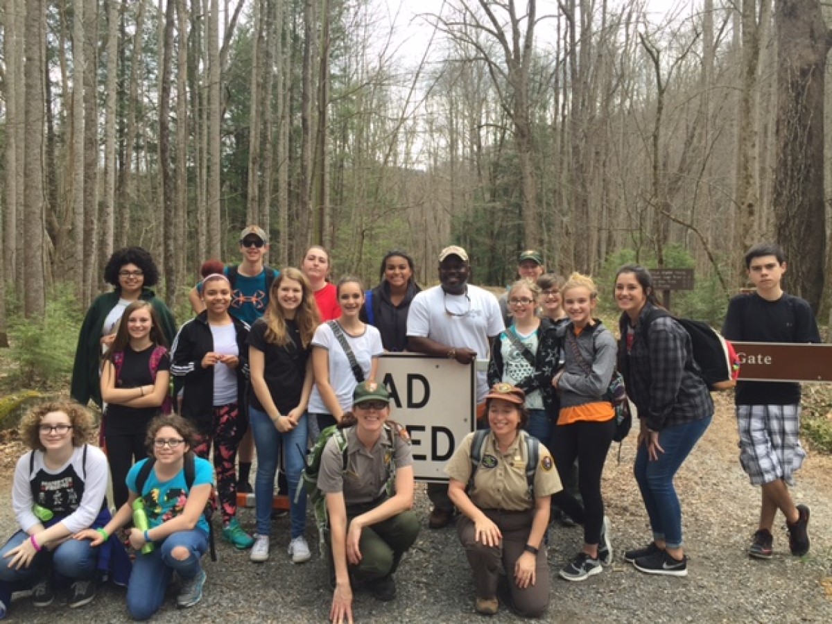 A large group of young people pose with kneeling rangers and park staff next to a road sign on a wide gravel path lined with skinny, bare trees.