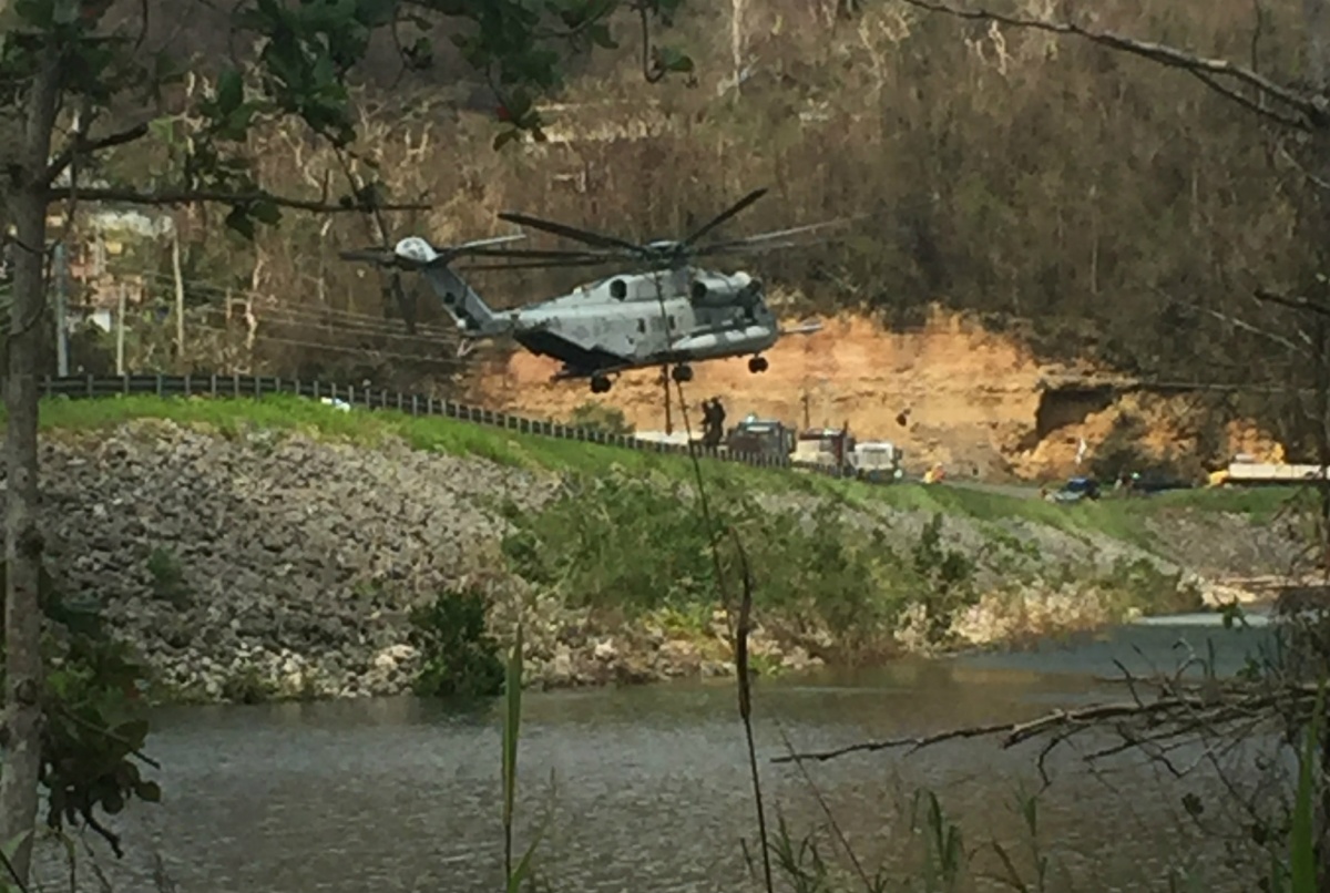 A large helicopter flies lower over a waterway next to a road.