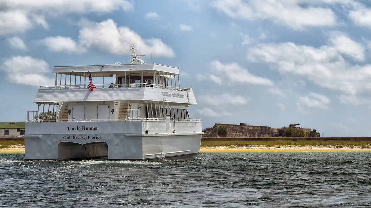 A large white boat travels across the water towards an old fort on the beach.
