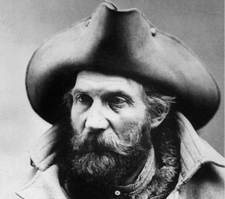 A bearded, rugged-looking man with a wide hat and warm coat looks off into the middle distance.