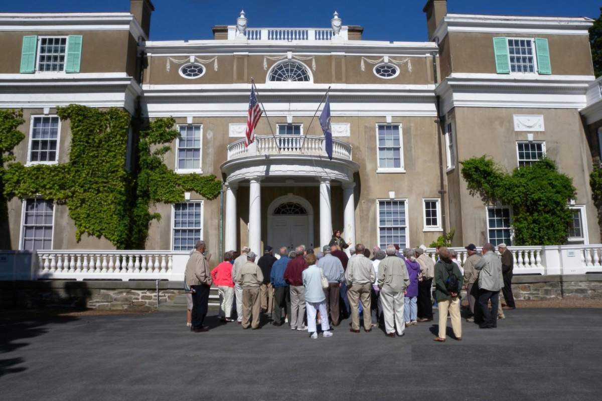 A small group of people stands outside a historic, three story mansion listening to a park ranger speaking.