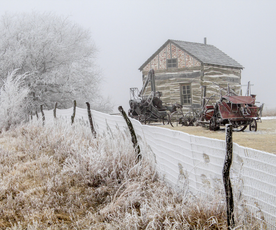 A wood and brick rustic house sits among a short leaning fence, white frostbitten shrubs and aging farming equipment.