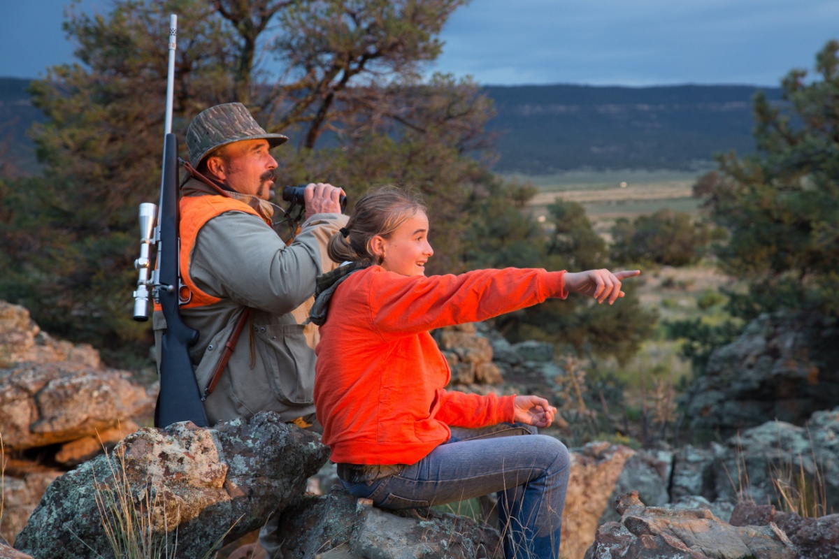 A child points at something as her dad wearing hunting gear looks through binoculars 