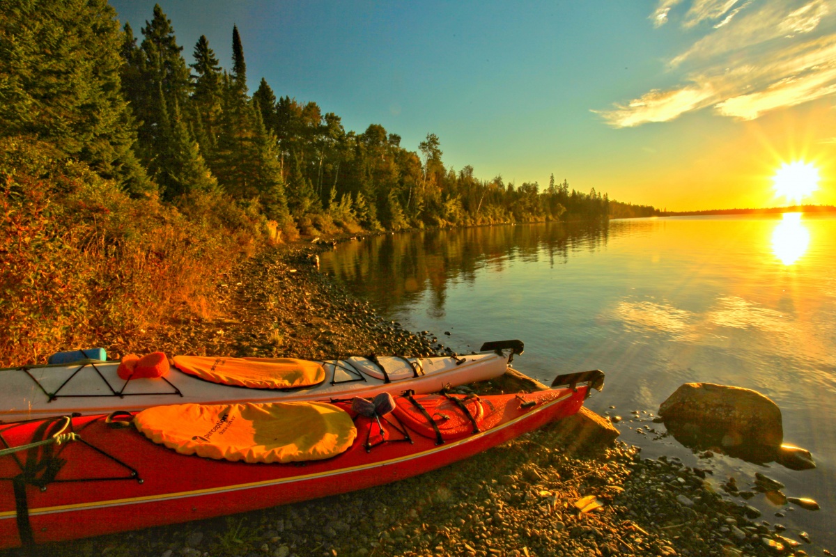 Two kayaks sit on the rocky shore. A bright sunset shines over the blue water, and a forest lines the shore's edge.