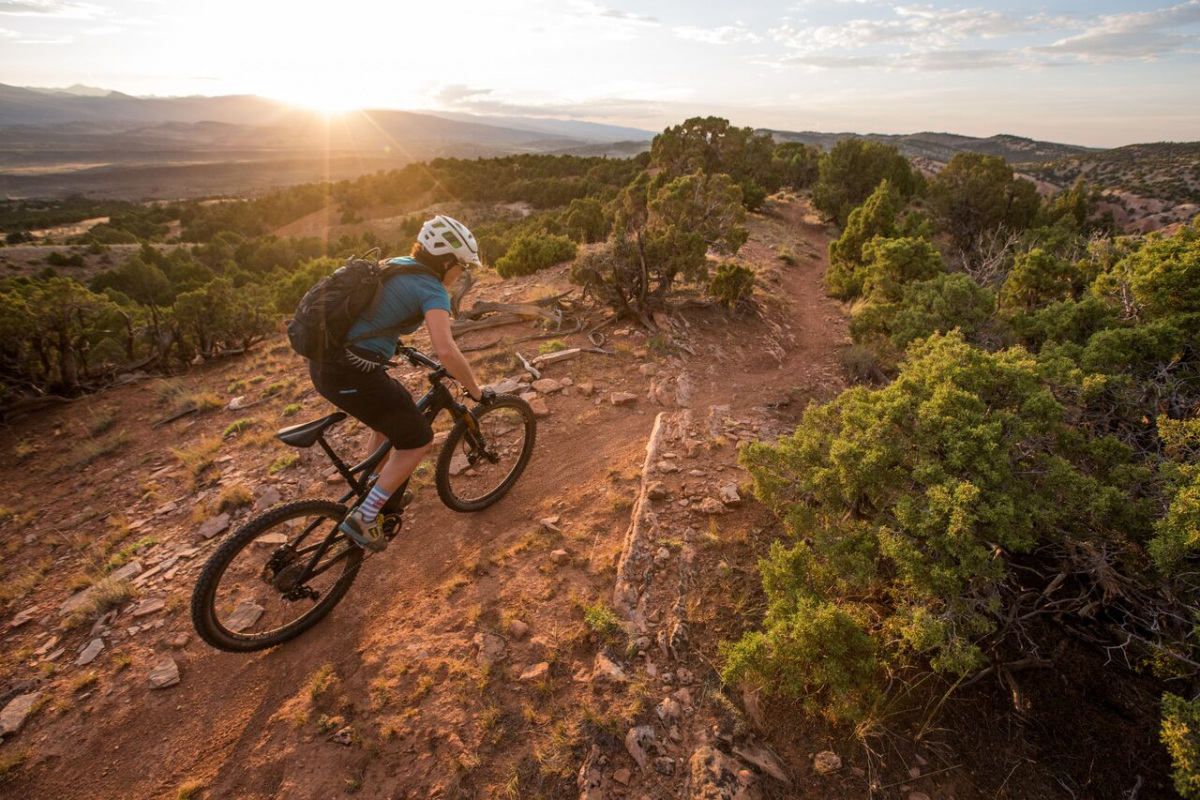 A white woman rides a mountain bike on a dirt trail along a rocky hilltop.