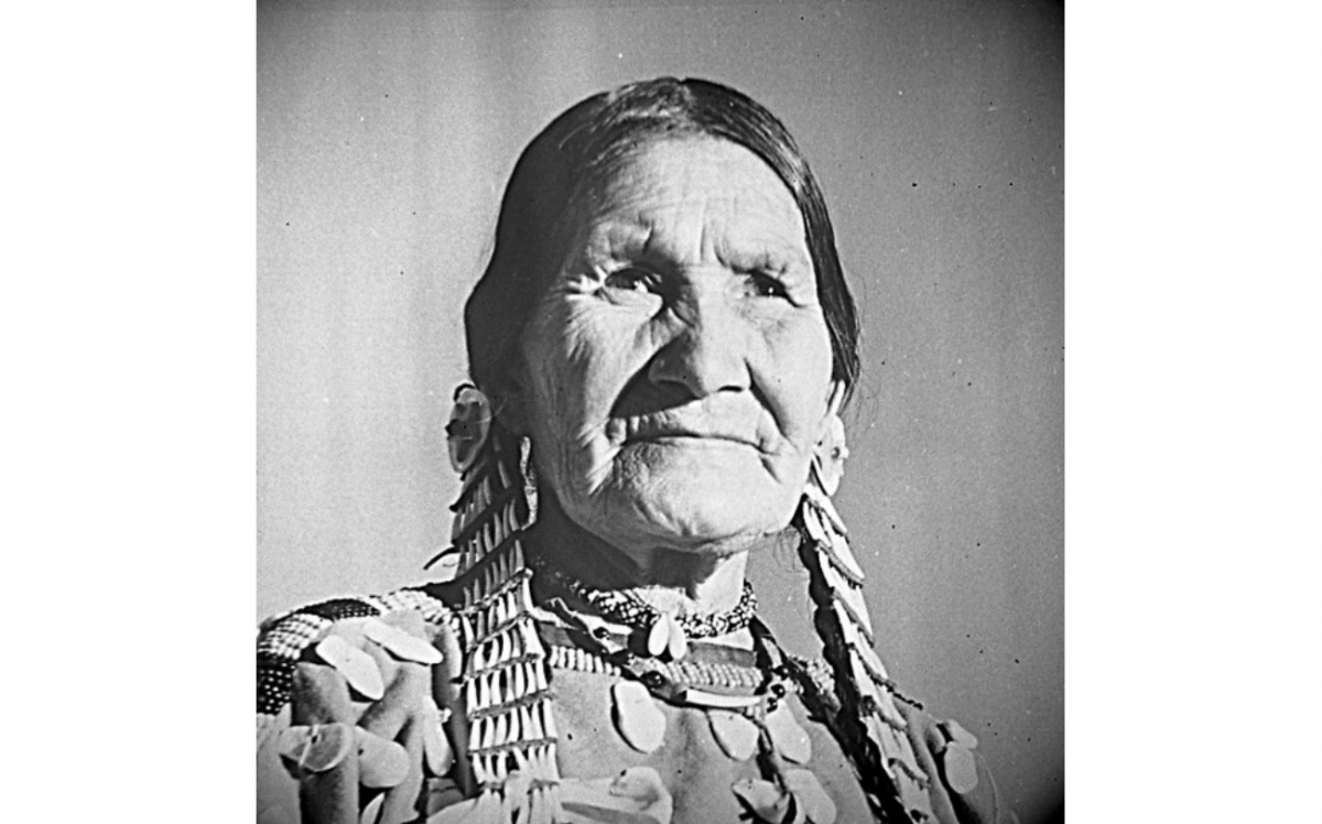 A woman, Julia Wades in the Water, stands up for this whole body shot. She is wearing traditional tribal dress and the photo is in black and white.