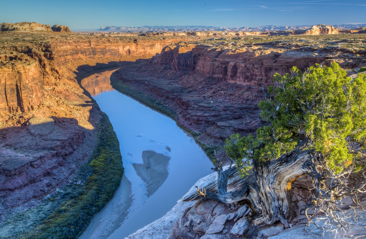 A flat blue river runs through a low, red rock canyon under a blue sky.