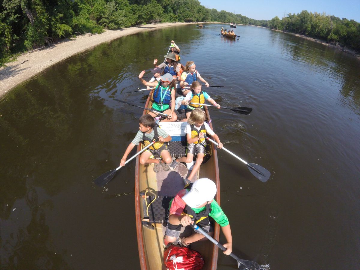 Children paddle in canoes along a muddy waterway.