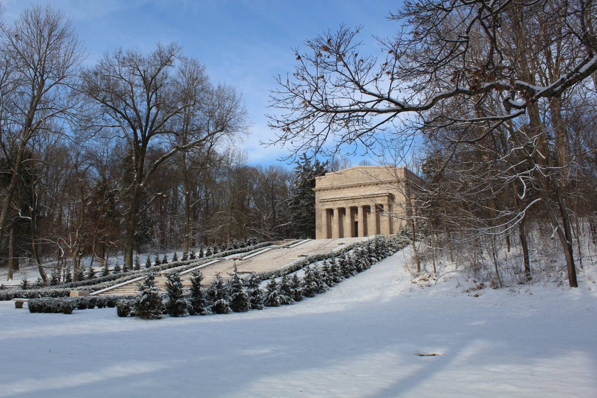 A large stone memorial stands at the top of a snowy hill with snow covered trees surrounding the path leading up to it.