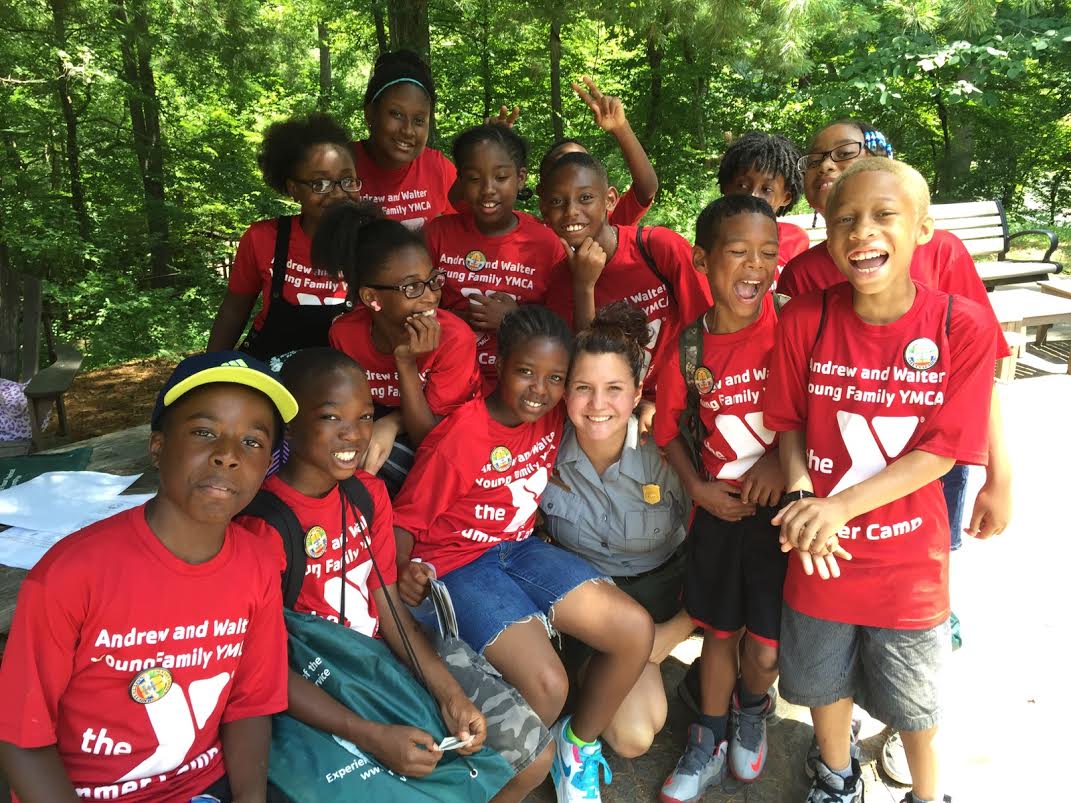 Children smile while posing with a NPS ranger outdoors.